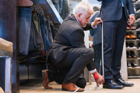 A tailor in a suit kneels on the floor, measuring the inseam of a client’s trousers with a measuring tape in a clothing store. Racks of pants and shelves of shoes are visible around them.
