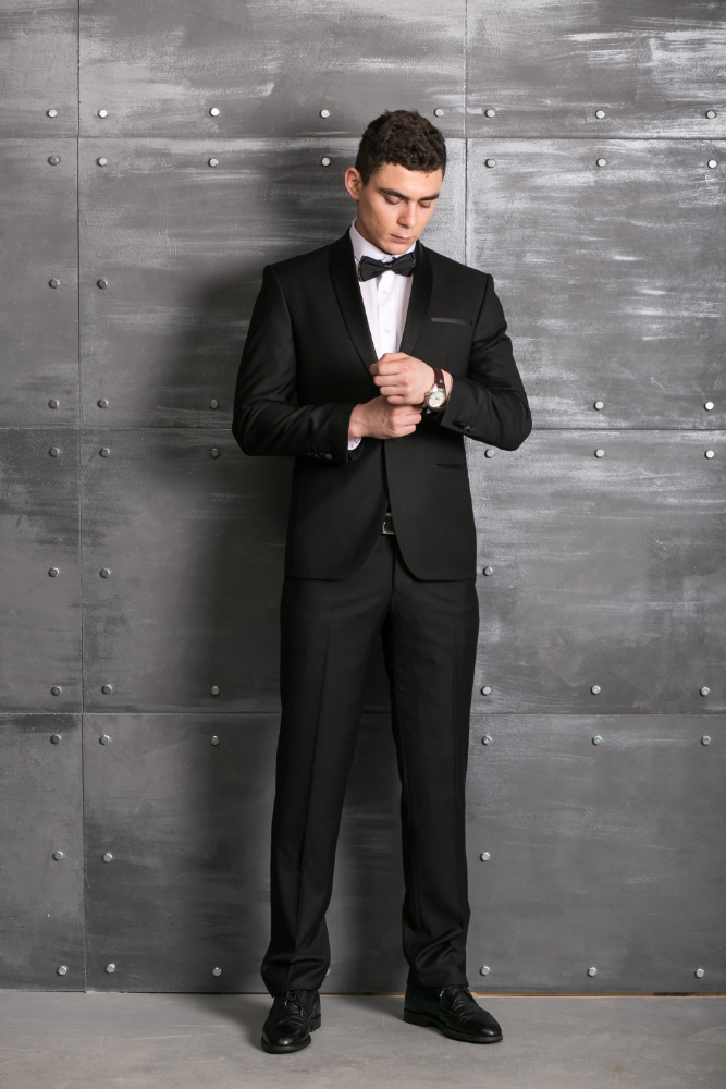 A man in a black tuxedo stands against a gray industrial-style wall with metal rivets, looking down while adjusting his cufflink.