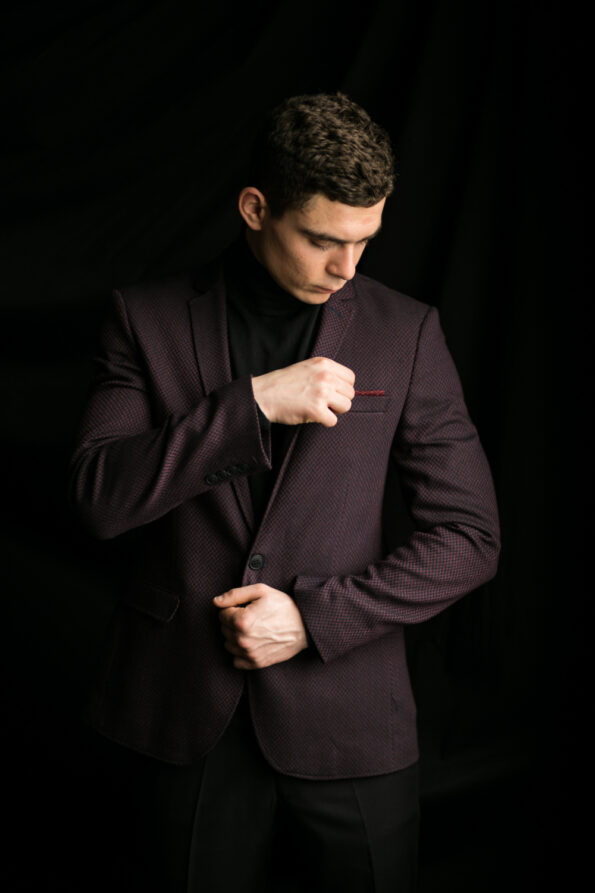 A man in a dark suit jacket and black turtleneck adjusts his sleeve while looking down, standing against a black background.